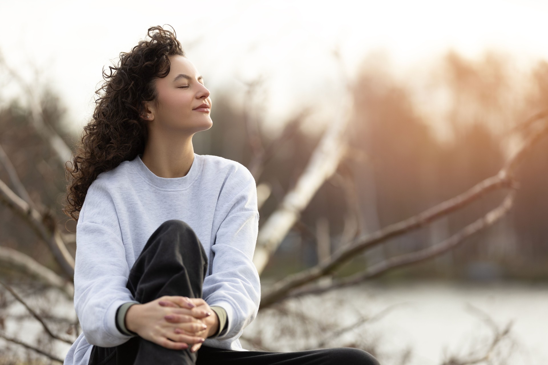 A beautiful girl with dark curly hair relaxing in sun and sitting on a fallen tree